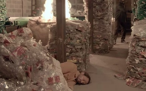 O ator Vladimir Brichta caracterizado como o Davi caído no chão, desacordado, em meio a pilhas de lixo que pegam fogo em cena de Amor de Mãe