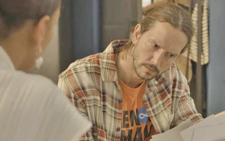 O ator Vladimir Brichta, com uma camisa xadrez sobre uma blusa laranja, lê um documento entregue por Taís Araújo, de costas no canto esquerdo. Ambos caracterizados como Davi e VItória em Amor de Mãe