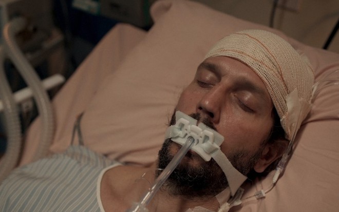 O ator Vladimir Brichta como Davi está deitado em uma cama de hospital, com bandanas ao redor da cabeça e um tubo ligado à boca em cena de Amor de Mãe