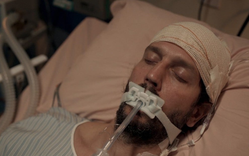 O ator Vladimir Brichta como Davi está deitado em uma cama de hospital, com bandanas ao redor da cabeça e um tubo ligado à boca em cena de Amor de Mãe