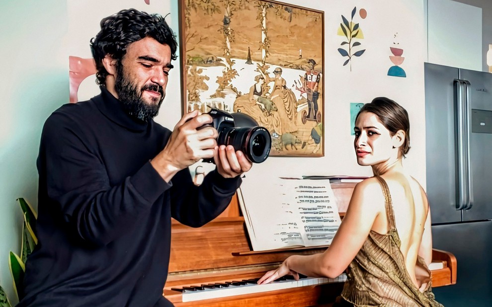 Caio Blat caracterizado como Manoel olha uma câmera fotográfica profissional enquanto Luisa Arraes, a Teresa, toca piano e vira o corpo para observar o ator com desprezo em cena de Amor & Sorte
