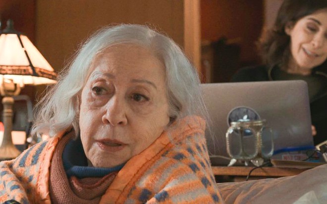 A atriz Fernanda Montenegro sentada em um sofá caracterizada como Gilda com Fernanda Torres, a Lúcia, ao fundo em um computador em cena de Amor & Sorte