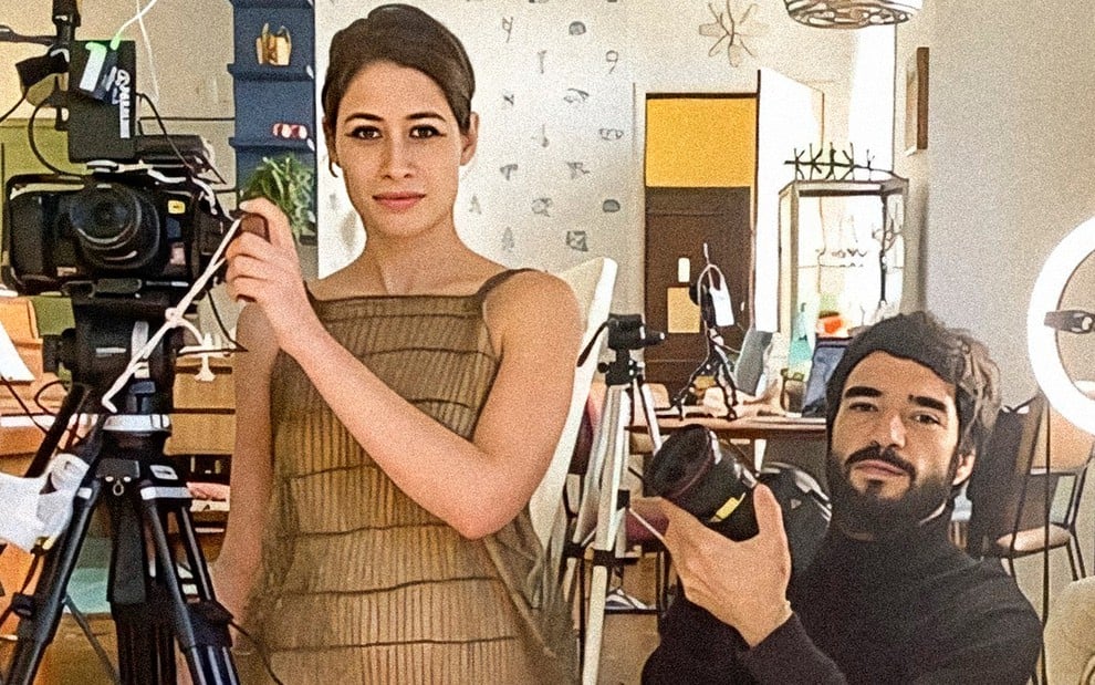 Em meio a equipamentos de filmagem, Luisa Arraes está de pé ao lado de uma câmera e Caio Blat está ajoelhado com uma câmera fotográfica nas mãos