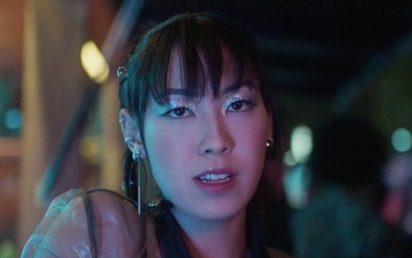 Ana Hikari em cena de As Five: caracterizada como Tina, personagem olha para frente com expressão séria