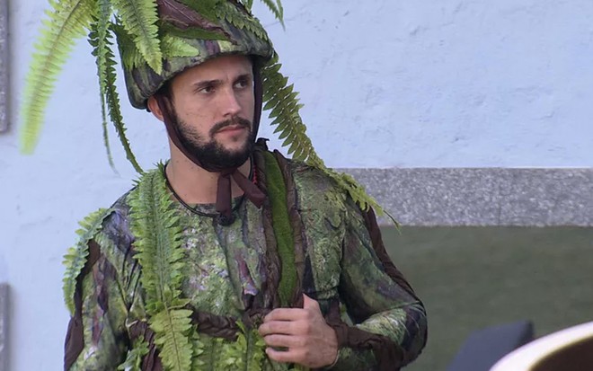 O preparador físico Arthur Picoli vestido como um chapéu com folhas de samambaia e uma roupa fechada com outras folhagens no jardim do BBB21