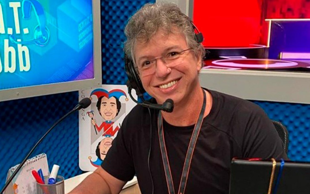 Boninho no Big Brother Brasil 20: sentado, diretor usa camiseta preta, cordão de crachá no pescoço, fone com microfone acoplado ao lado de seu rosto