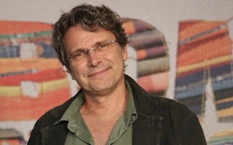 O diretor Luiz Henrique Rios, com uma blusa verde e casaco preto, de óculos, olha para a câmera em frente a um backdrop com a logo de Bom Sucesso