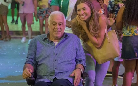 Antonio Fagundes com camiseta azul sentado na cadeira de rodas é conduzido por Grazi Massafera em cena da novela Bom Sucesso