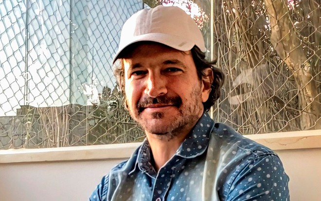 O ator Caco Cicoler com boné, de bigode, posa em frente a uma janela com rede de proteção