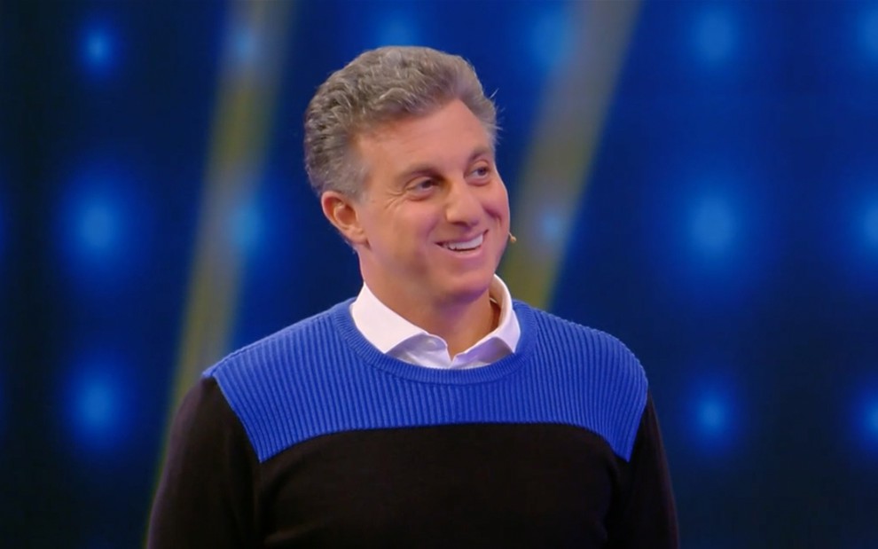 O apresentador Luciano Huck com um suéter listrado azul e preto com um sorriso amarelo no rosto no palco do Caldeirão