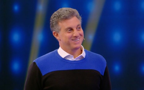 O apresentador Luciano Huck com um suéter listrado azul e preto com um sorriso amarelo no rosto no palco do Caldeirão