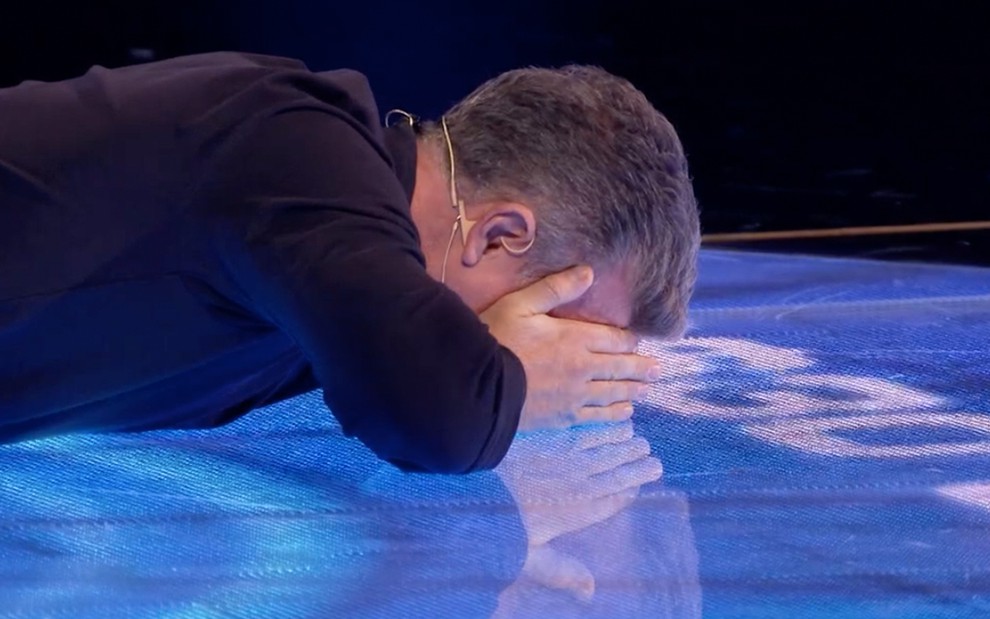 O apresentador Luciano Huck com uma blusa preta de manga comprida está deitado no chão com a mão cobrindo os olhos no palco do The Wall, no Caldeirão do Huck