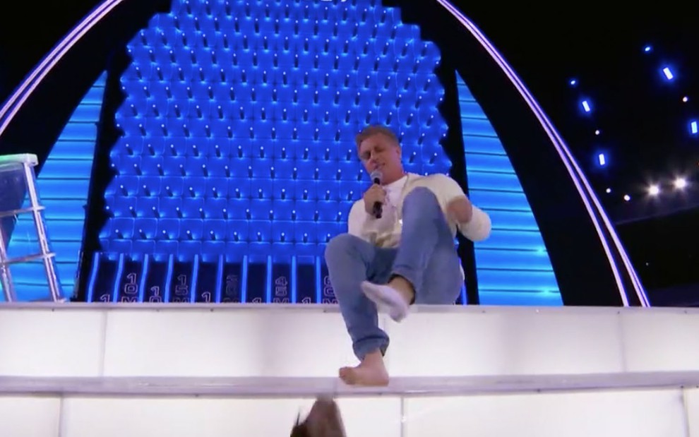 O apresentador Luciano Huck sentado no palco tirando os sapatos com um microfone na mão direita 