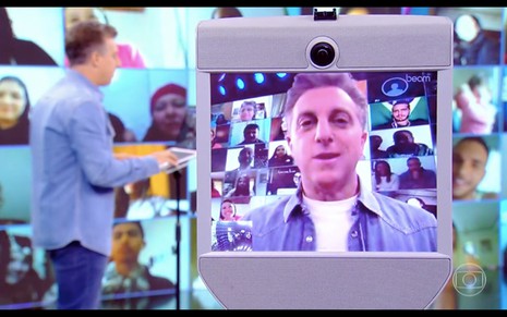 Luciano Huck ao fundo comanda um robô que mostra o seu rosto em uma tela