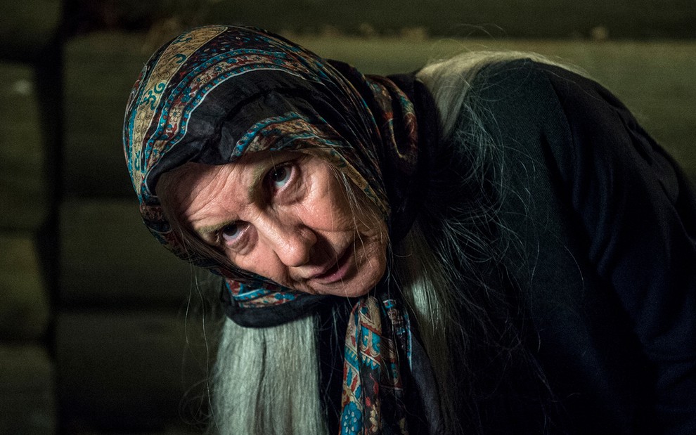 A atriz Cássia Kis com um véu colorido na cabeça e longos cabelos grisalhos, se curva em direção ao chão como a bruxa Haia em Desalma