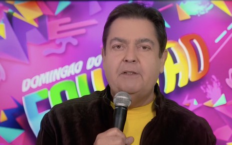 Fausto Silva com microfone na mão na frente de um backdrop com os dizeres Domingão do Faustão