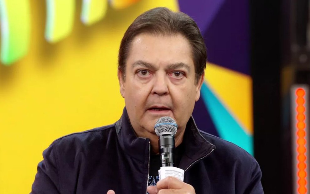 O apresentador Fausto Silva no palco do Domingão do Faustão com um microfone na mão direta e uma jaqueta azul-marinho