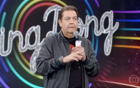 O apresentador Fausto Silva no palco do Domingão do Faustão com a logo do Ding Dong ao fundo