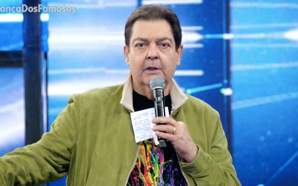 O apresentador Fausto Silva com uma blusa preta e um casaco verde, ele segura o microfone com a mão direita