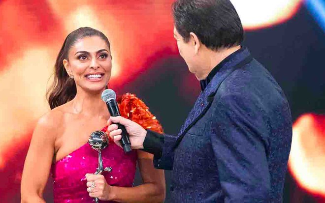 A atriz Juliana Paes segurando uma estatueta, sorri enquanto fala no microfone estendido por Fausto Silva, de costas