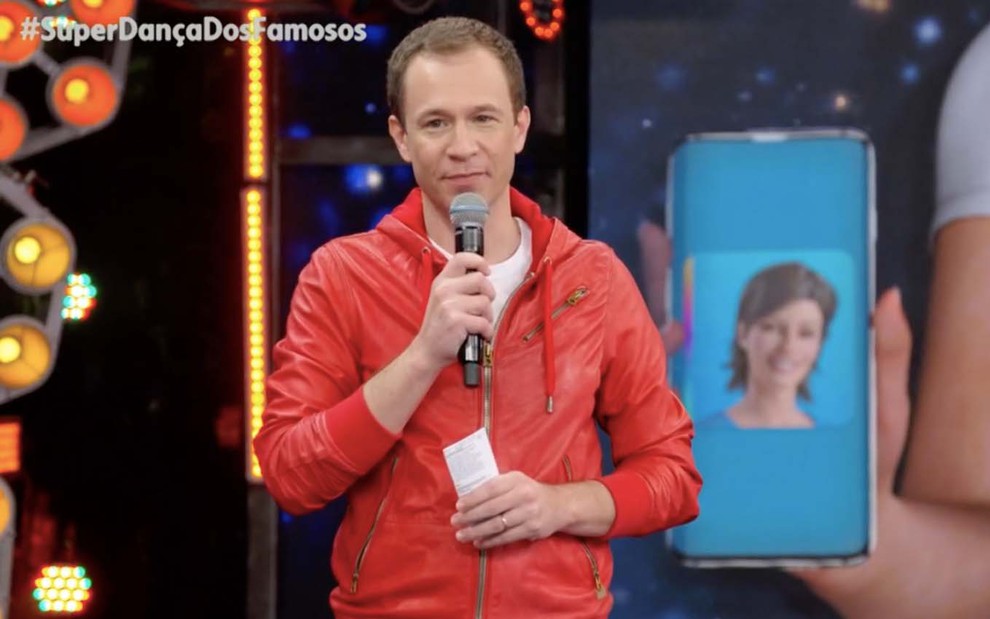 O jornalista Tiago Leifert no palco do Domingão do Faustão com um casaco vermelho, uma blusa branca e um microfone na mão direita