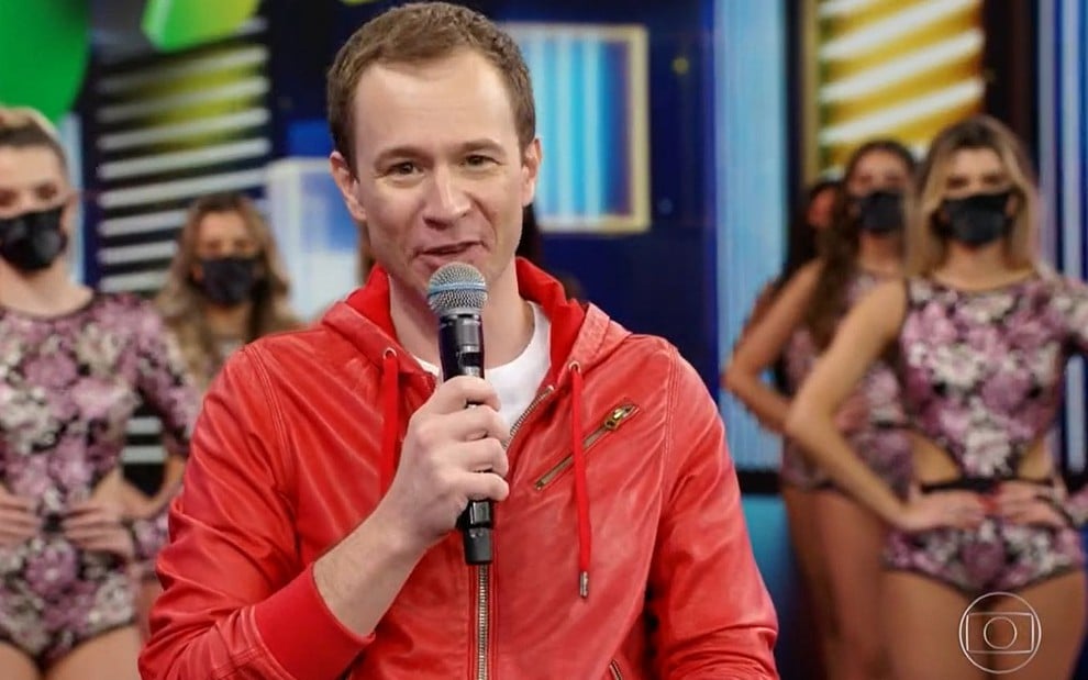 O jornalista Tiago Leifert com um casaco vermelho e uma blusa branca no palco do Domingão, com um microfone na mão esquerda