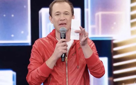 O apresentador Tiago Leifert segura um microfone com a mão direita e uma ficha com a mão esquerda, ele está com um casaco vermelho e uma blusa branca