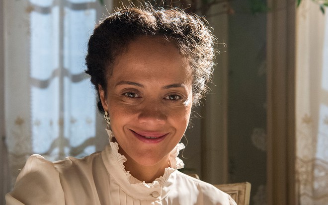 A atriz Aline Borges, com um vestido branco e cabelos presos, sorri diretamente para a câmera caracterizada como a Selma em cena de Éramos Seis