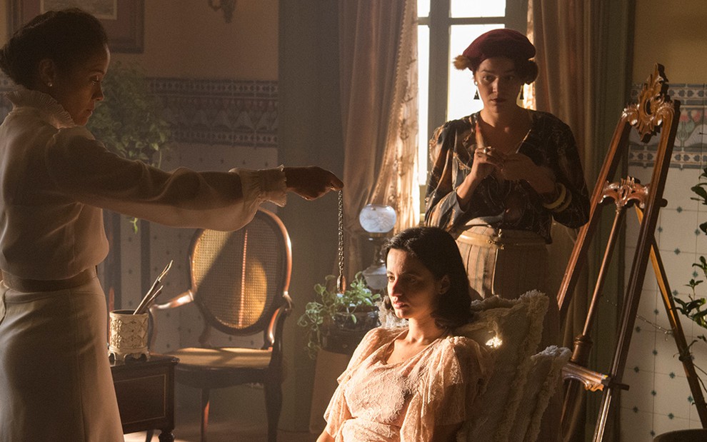 Aline Borges, caracterizada como selma, segura um pêndulo diante de Júlia Stockler, a Justina, que está sentada em uma cadeira a frente de Joana de Verona, a Adelaide, em cena de Éramos Sesi