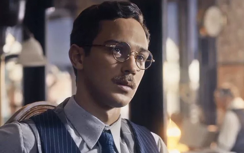 O ator André Luiz Frambach com colete, grava e óculos, olha para a direita, caracterizado como Julinho em cena de Éramos Seis
