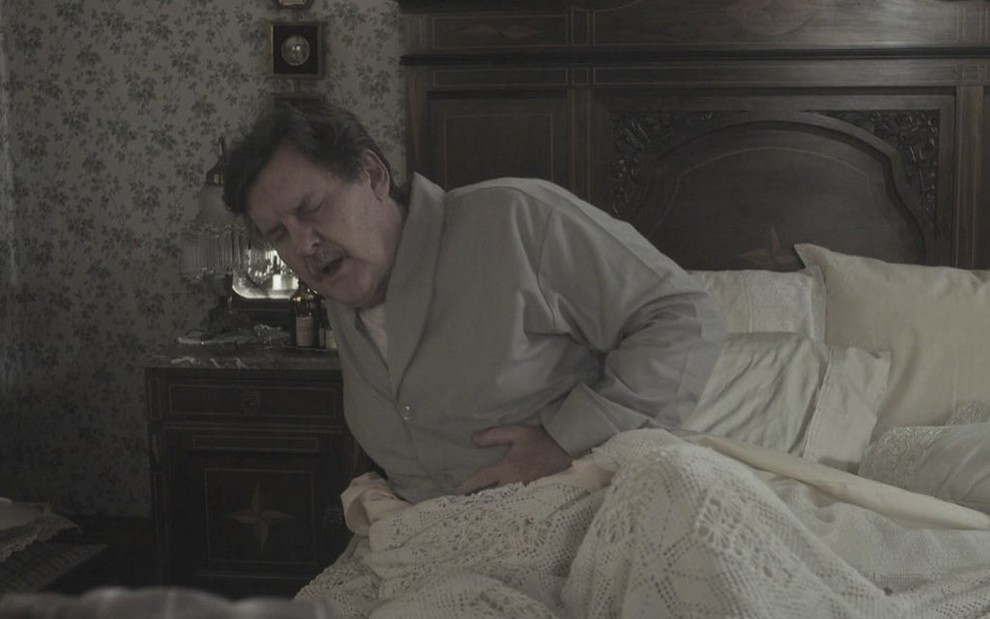 O ator Antonio Calloni, caracterizado como Júlio em Éramos Seis, coloca a mão na barriga como se sentisse uma forte dor, ele está acamado