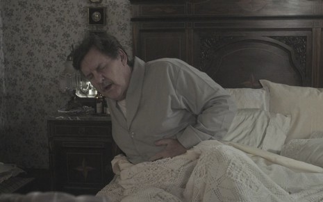 O ator Antonio Calloni, caracterizado como Júlio em Éramos Seis, coloca a mão na barriga como se sentisse uma forte dor, ele está acamado