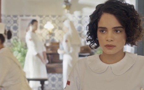 A atriz Carol Macedo, com um uniforme branco de enfermeira, caracterizada como a Inês em cena de Éramos Seis