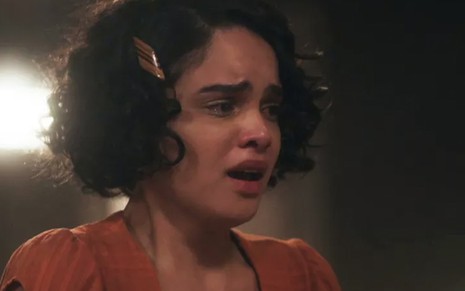 A atriz Carol Macedo olha para a direita, com lágrimas nos olhos e vestido vermelho, caracterizada como a Inês em cena de Éramos Seis