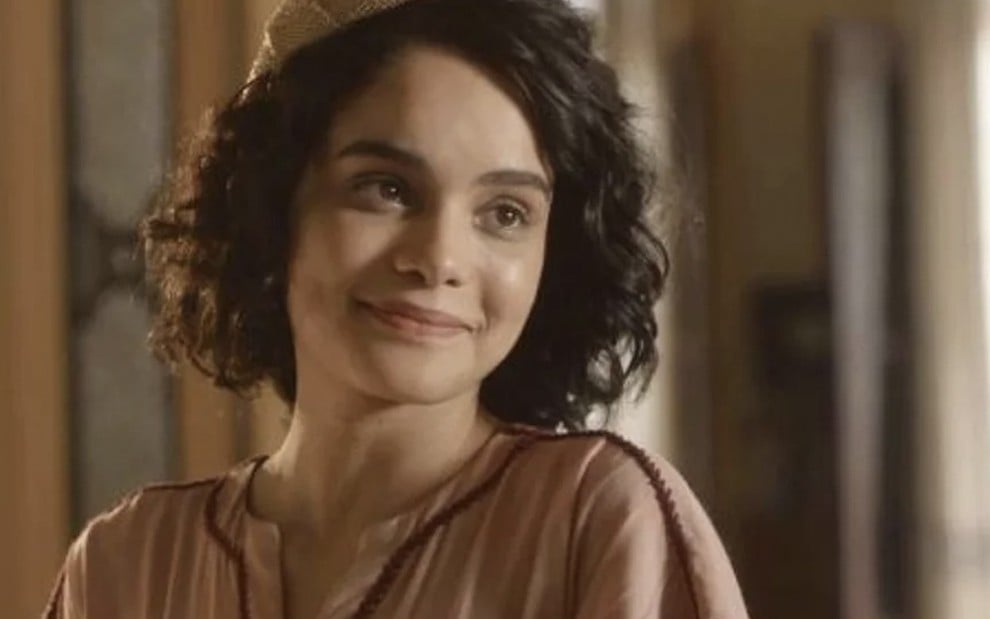 A atriz Carol Macedo, com uma roupa bege, sorri com o rosto levemente inclinado para a direita caracterizada como a Inês em cena de Éramos Seis