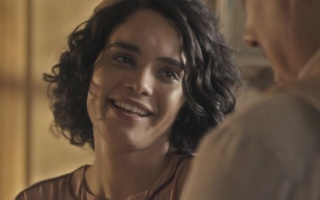 A atriz Carol Macedo olha para Cássio Gabus Mendes, de costas, e sorri caracterizada como a Inês em cena de Éramos Seis