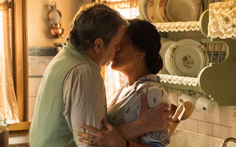Cássio Gabus Mendes e Gloria Pires, caracterizados como Afonso e Lola, se beijam em uma cozinha em cena de Éramos Seis