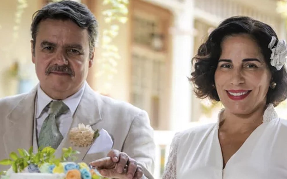 O ator Cássio Gabus Mendes e a atriz Gloria Pires, nesta ordem, cortam um bolo vestidos de branco durante cena de casamento de Afonso e Lola em Éramos Seis