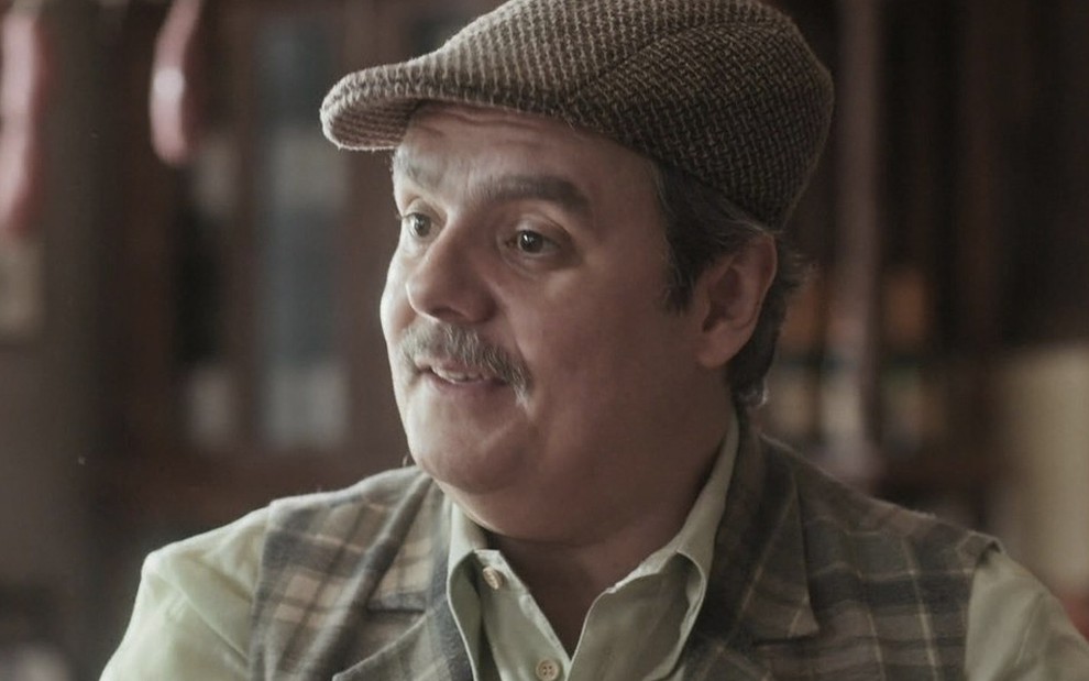 O ator Cássio Gabus Mendes, com uma boina marrom, sorri enquanto olha para a esquerda caracterizado como o Afonso em cena de Éramos Seis