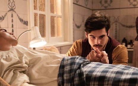 Danilo Mesquita, o Carlos, deitado em uma cama de hospital, segurando a mão de Nicolas Prattes, o Alfredo, em cena de Éramos Seis