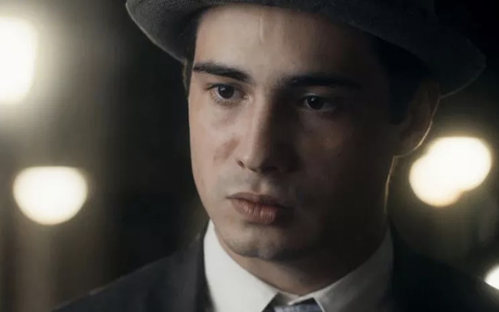 O ator Danilo Mesquita, em um close de terno preto e chapéu, caracterizado como Carlos em cena noturna de Éramos Seis
