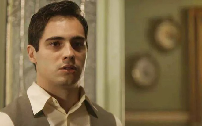 O ator Danilo Mesquita, com roupa social e colete, olha para a direita, caracterizado como o Carlos em cena de Éramos Seis