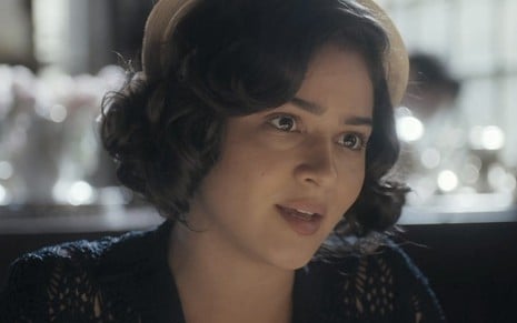 A atriz Giullia Buscacio, com um chapéu bege e uma roupa preta, olha para a direta caracterizada como a Isabel em cena de Éramos Seis