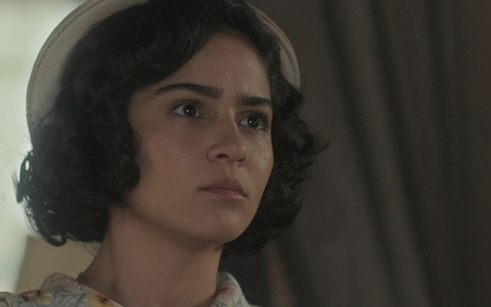 A atriz Giullia Buscacio, com um pequeno chapéu, olha para o lado direito, caracterizada como a Isabel em cena de Éramos Seis