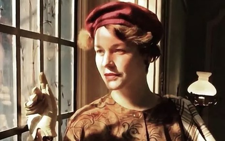 A atriz Joana de Verona olha por uma janela, em meio a raios de sol, caracterizada como a Adelaide em cena de Éramos Seis