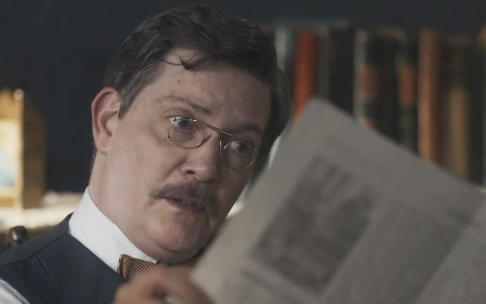 O ator Kiko Mascarenhas, com um óculos redondo, com uma armação final, lê um jornal boquiaberto, caracterizado como o Virgulino de Éramos Seis