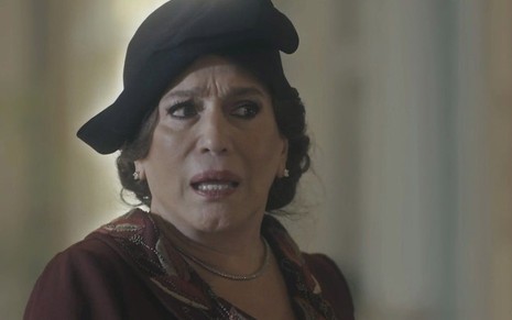 A atriz Susana Vieira caracterizada como a personagem Emília, de chapéu preto e vestido escuro, olha com surpresa em cena de Éramos Seis