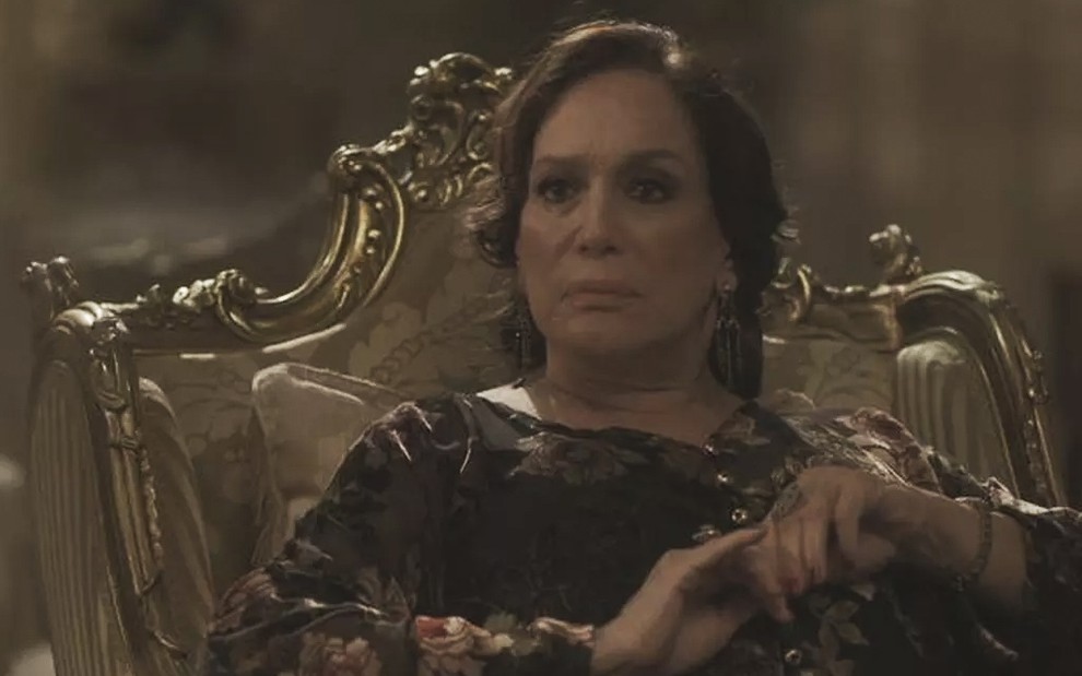 A atriz Susana Vieira, com um vestido preto, sentado em uma cadeira dourada, caracterizada como a Emília em cena de Éramos Seis