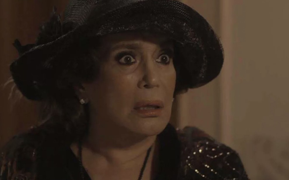 A atriz Susana Vieira, com um chapéu e vestido pretos, com expressão de horror, caracterizada como a Emília em cena de Éramos Seis