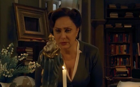 A atriz Eliane Giardini caracterizada como Anastácia se curva em frente a sua santa e uma vela em cena de Êta Mundo Bom!